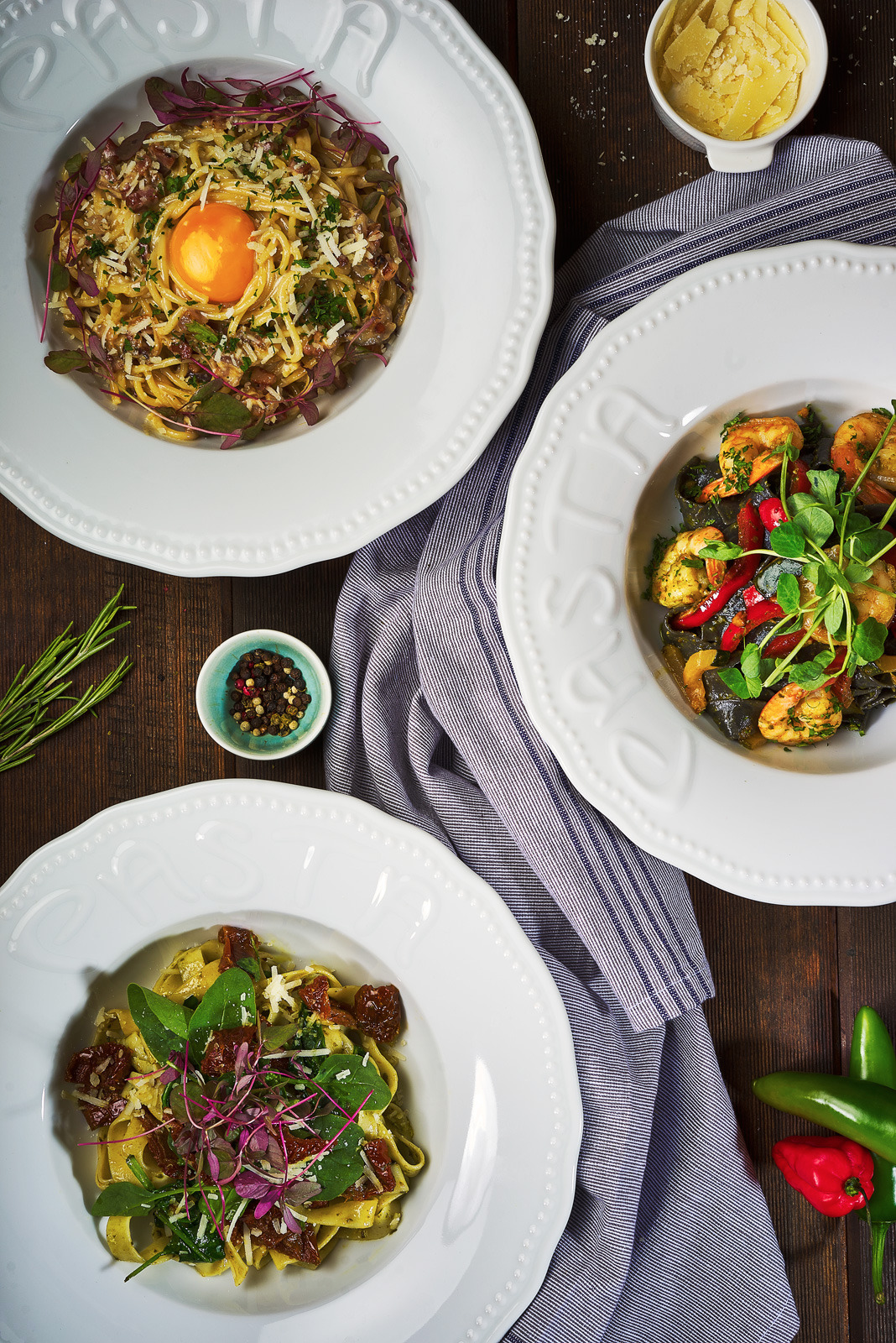 Trzy talerze z różnymi rodzajami makaronu na drewnianym stole, udekorowane ziołami i przyprawami, na tle lnianej ściereczki w paski.