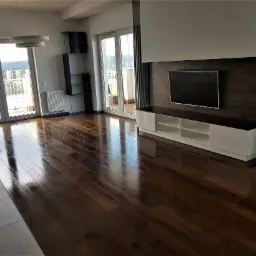 Wykończony salon z ciemnym, lakierowanym parkietem ułożonym w deski, odbijającym światło z okien balkonowych. Na ścianie wisi telewizor nad białą szafką RTV. Widok na miasto z balkonu.