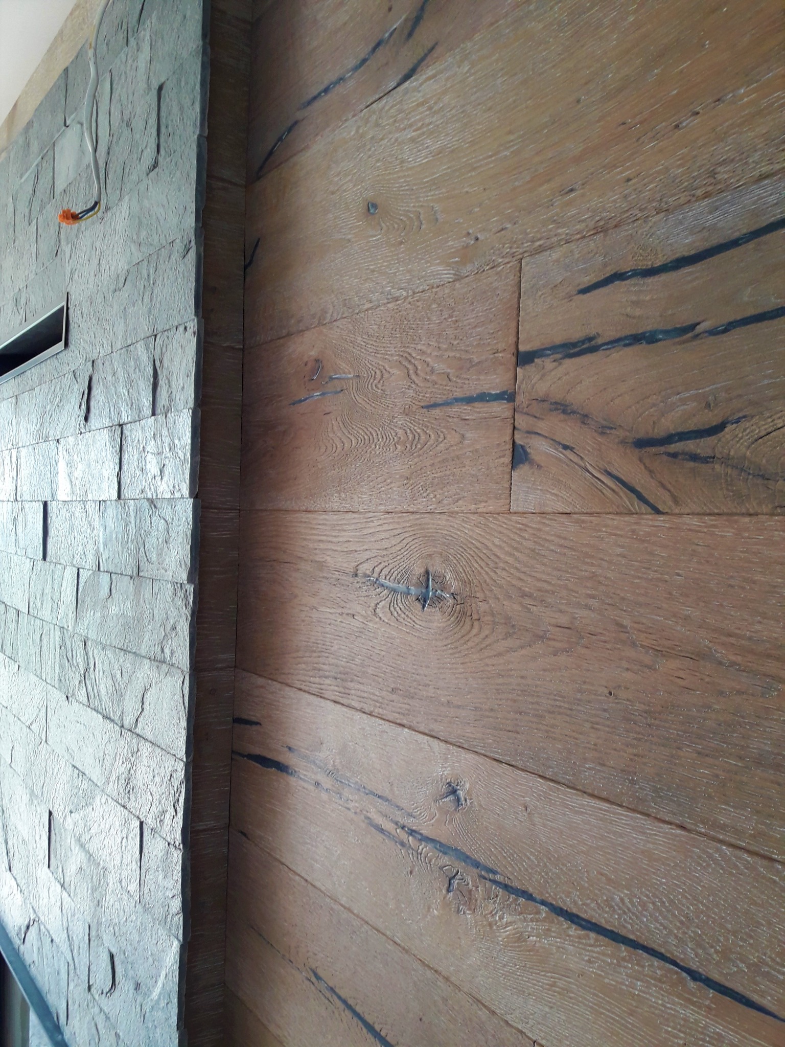 Wykończenie wnętrza: ściana z szarej, łupkowej okładziny kamiennej sąsiaduje z podłogą z parkietu z wyraźnym usłojeniem i czarnymi akcentami.