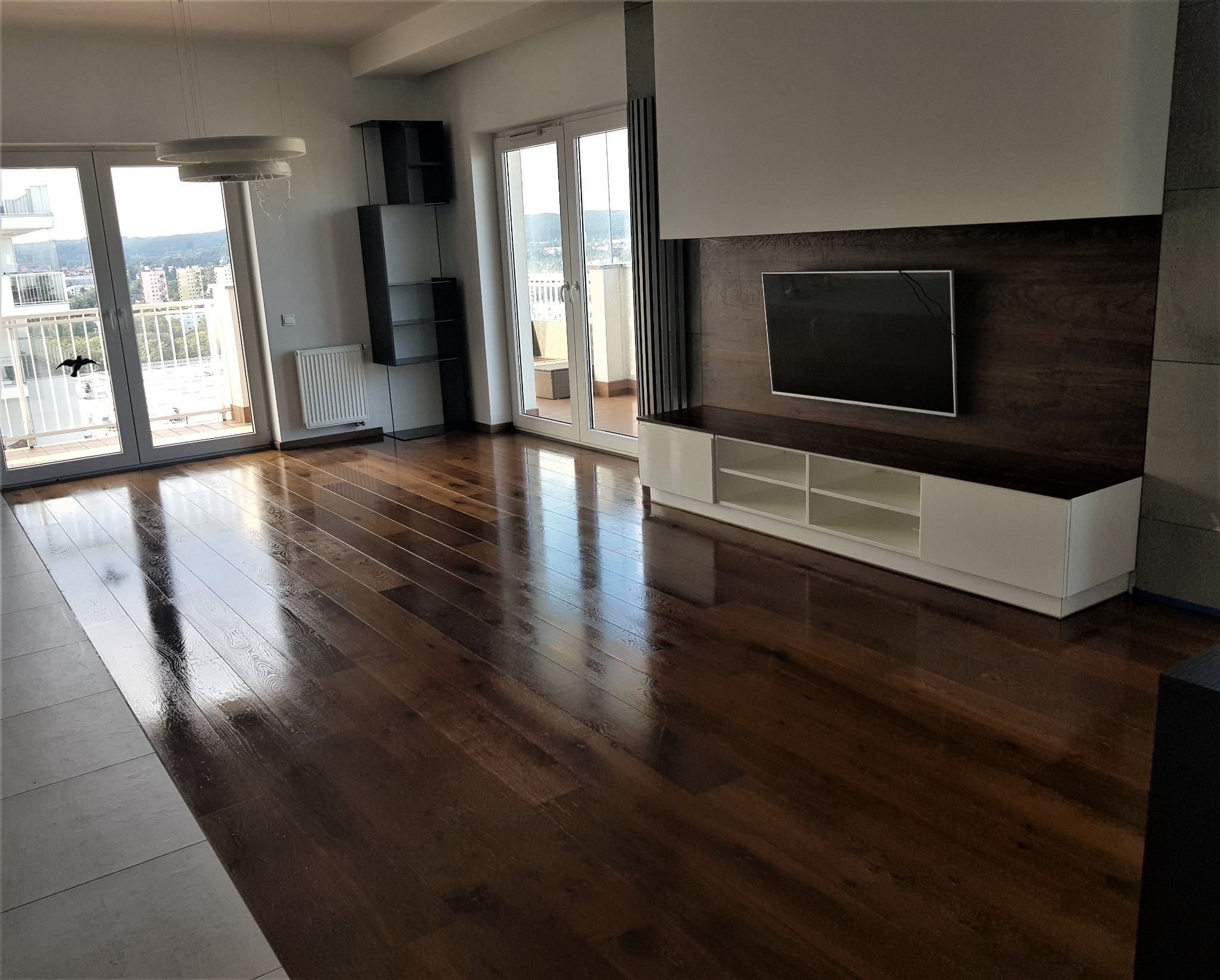 Wykończony salon z ciemnym, lakierowanym parkietem ułożonym w deski, odbijającym światło z okien balkonowych. Na ścianie wisi telewizor nad białą szafką RTV. Widok na miasto z balkonu.