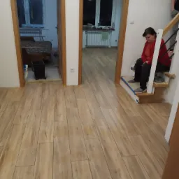 Układanie podłogi z paneli drewnopodobnych, widoczne dwa wejścia do pomieszczeń, kobieta siedzi na schodach w czerwonej bluzce.