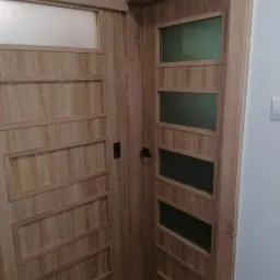 Dwa skrzydła drzwi wewnętrznych w odcieniach drewna: jedno z poziomymi prostokątnymi wgłębieniami, drugie z czterema prostokątnymi przeszkleniami, widoczne klamki.