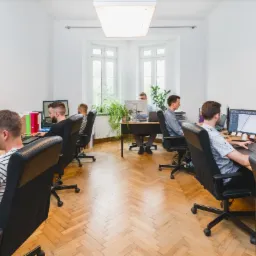 Jasne biuro z parkietem, pracownicy przy biurkach, na monitorach widoczne schematy i projekty.