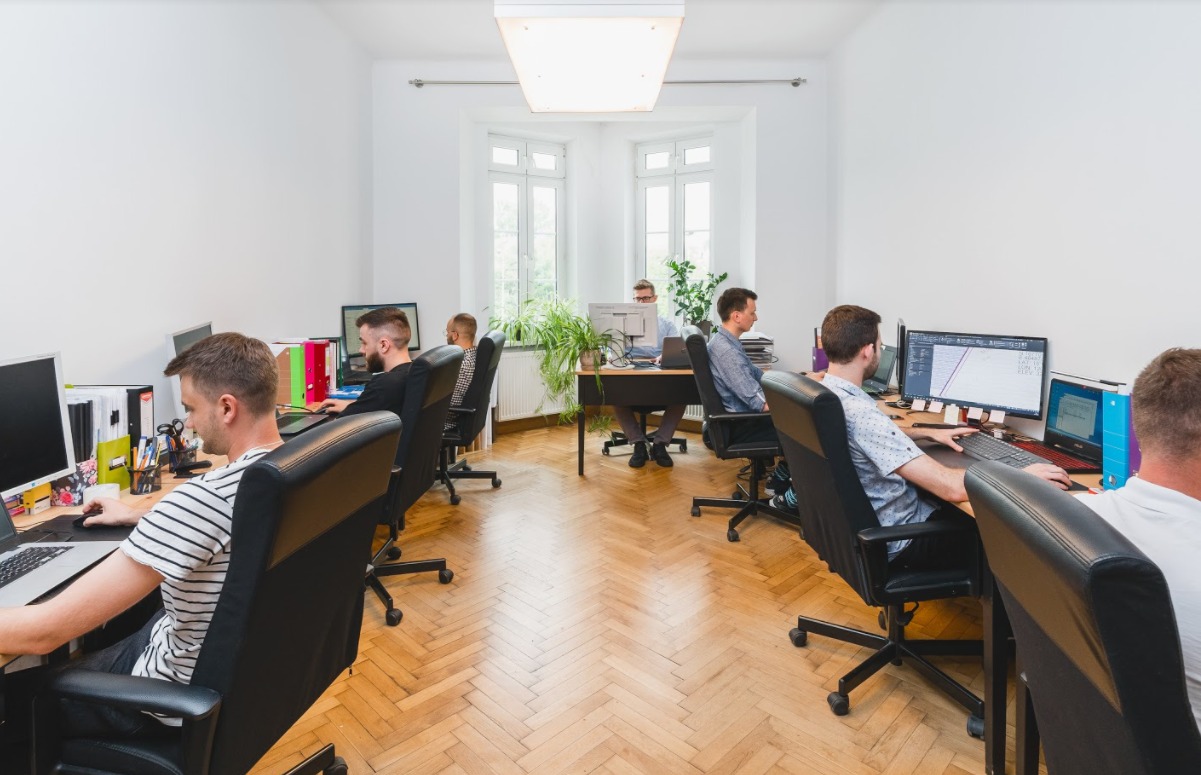Jasne biuro z parkietem, pracownicy przy biurkach, na monitorach widoczne schematy i projekty.