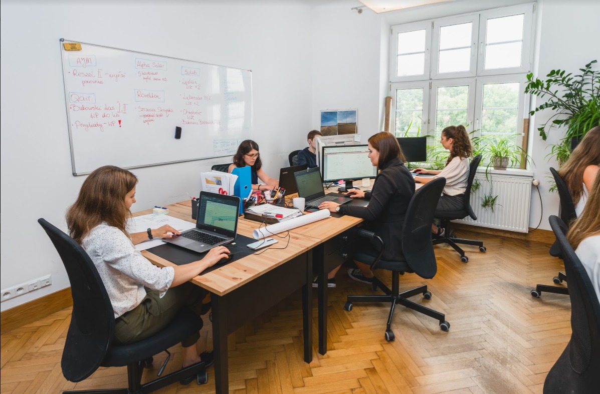 Biuro projektowe z pracownikami przy biurkach, widoczna tablica suchościeralna z zapiskami, plany instalacji elektrycznych na biurku.