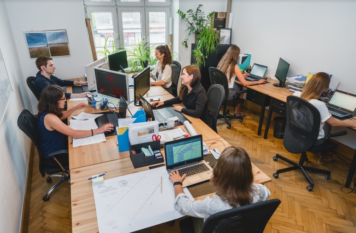 Biuro projektowe: zespół inżynierów pracuje nad projektami instalacji, widoczne komputery, plany na papierze, biurka z jasnego drewna, rośliny doniczkowe, duże okno z widokiem na zewnątrz.
