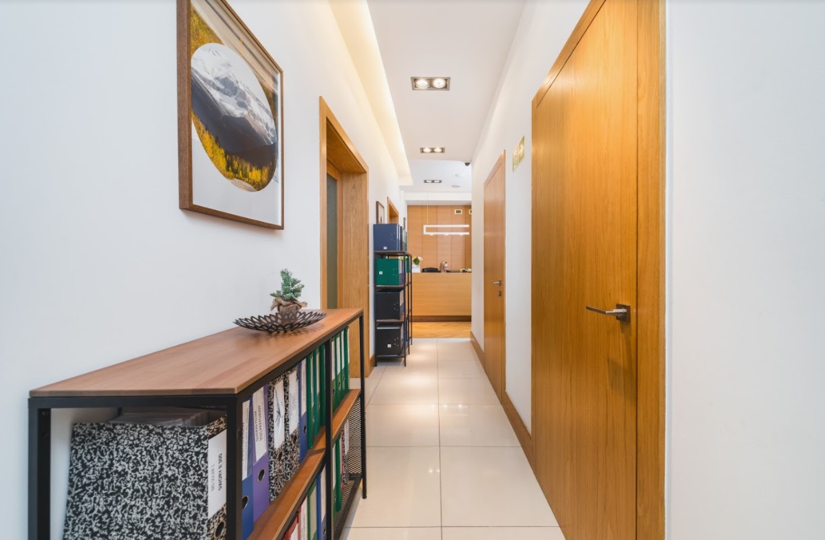Korytarz biurowy z białymi ścianami, drewnianymi drzwiami i regałem z segregatorami, zakończony recepcją z oświetleniem LED.