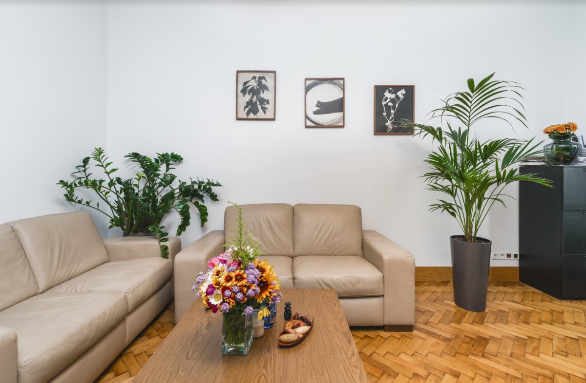 Jasne wnętrze biura z beżowymi sofami, drewnianym stolikiem z bukietem kwiatów, roślinami doniczkowymi i trzema obrazkami na ścianie