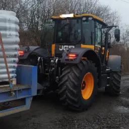 Ciągnik JCB z przyczepą załadowaną paletami z workami z ekogroszkiem w Łochowie, widok z tyłu, szare niebo w tle.