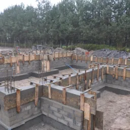 Budowa fundamentów domu jednorodzinnego z bloczków betonowych, szalunki drewniane, widoczne zbrojenia, w tle rząd drzew iglastych.