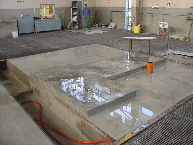 Świeżo wylana, mokra posadzka jastrychowa w hali przemysłowej, z widocznym poziomowaniem, pomarańczowym wiadrem i pracownikiem w tle.