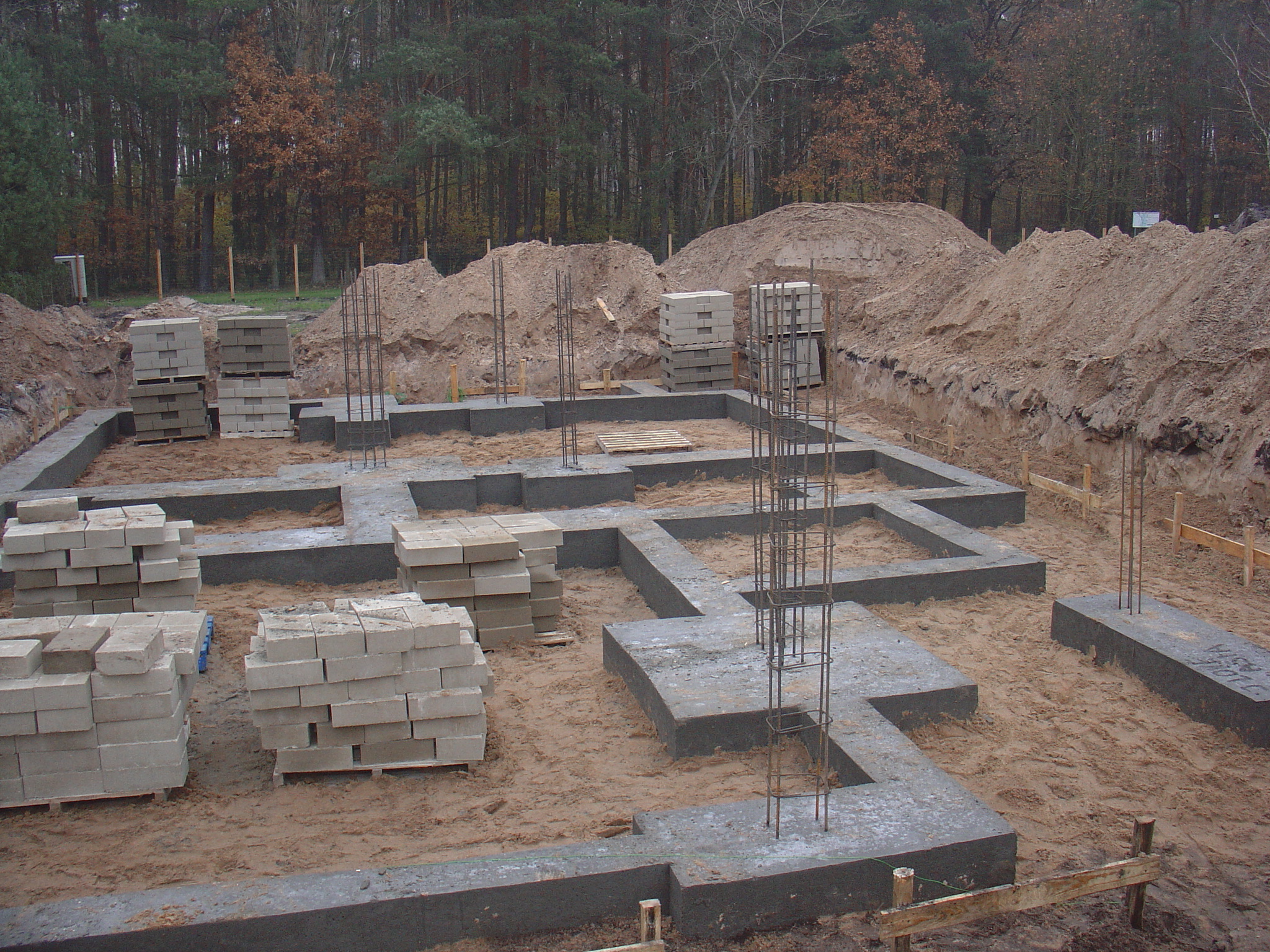 Wykopane fundamenty domu z betonowymi ławami, zbrojeniami i paletami z bloczkami betonowymi na piaszczystym podłożu, otoczone ziemią i drzewami w tle.