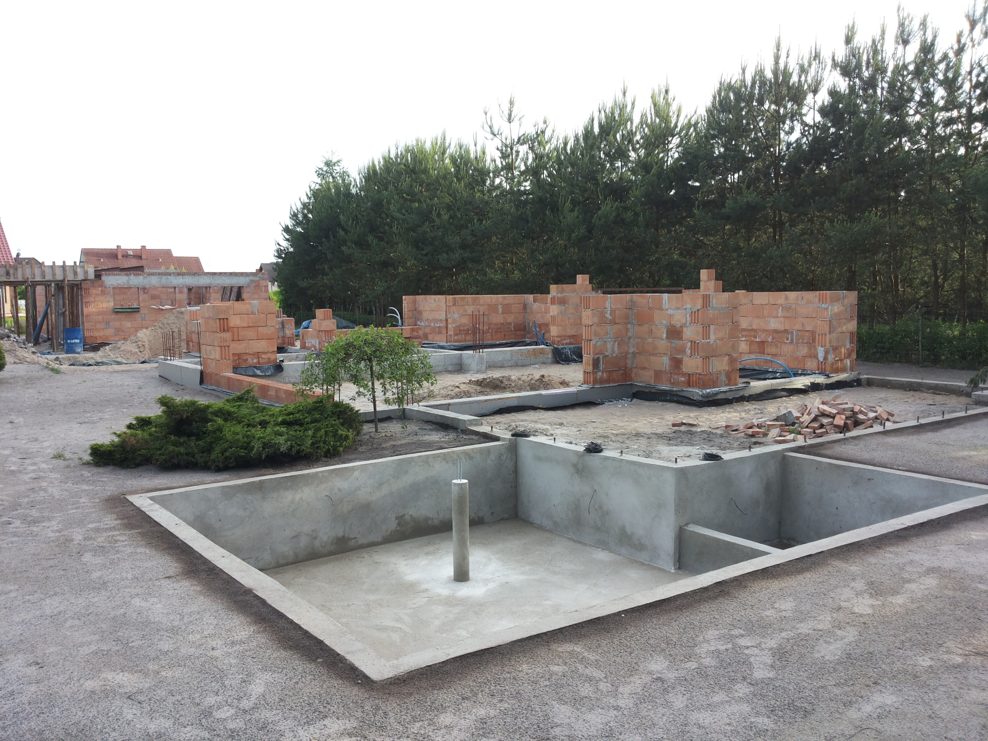 Fundamenty domu w trakcie budowy, widoczne ściany z cegły, betonowe wylewki, zieleń drzew w tle.