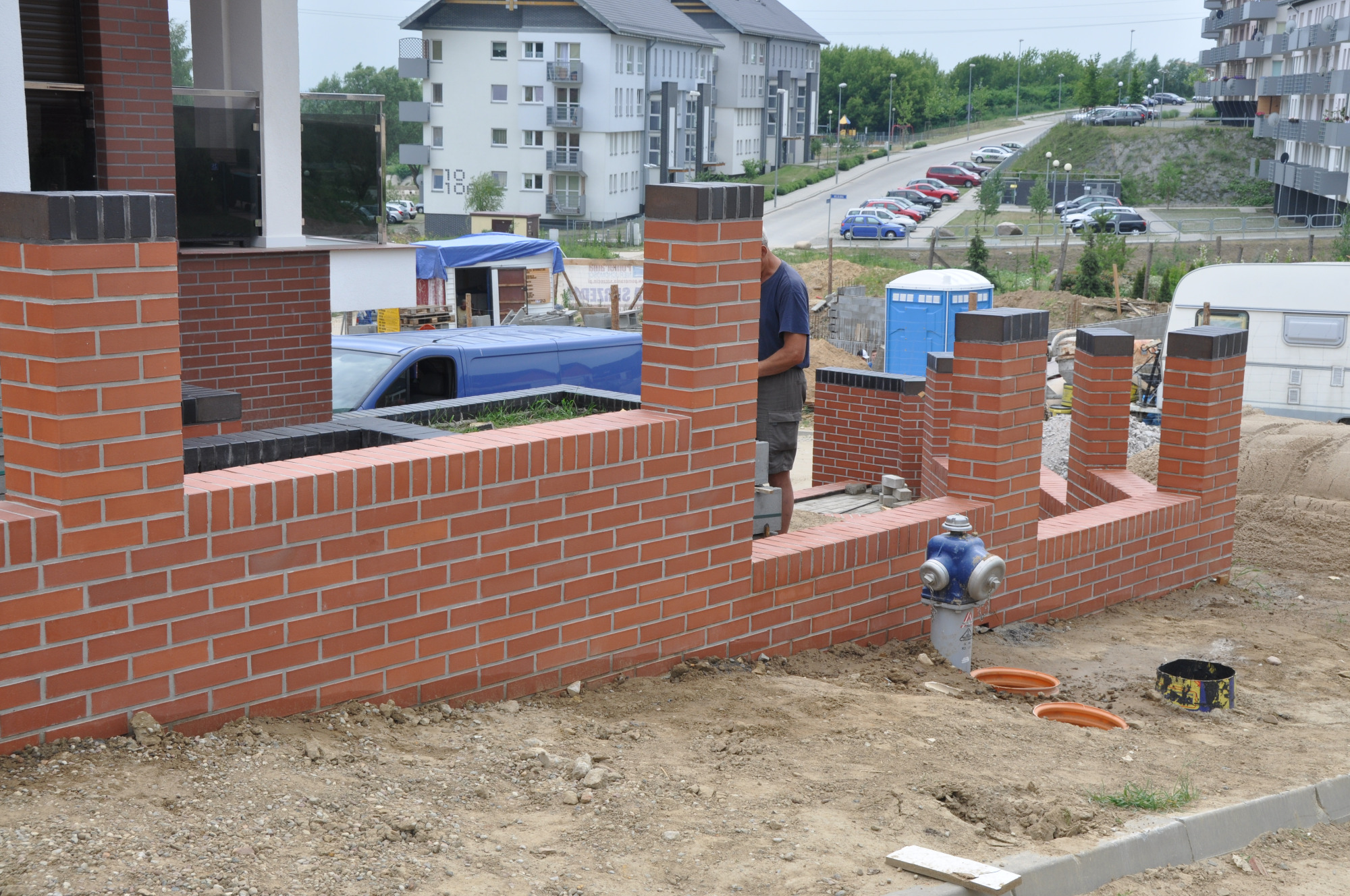 Wznoszenie muru z czerwonej cegły na terenie budowy z widocznym hydrantem, zaparkowanym samochodem dostawczym i budynkami mieszkalnymi w tle.