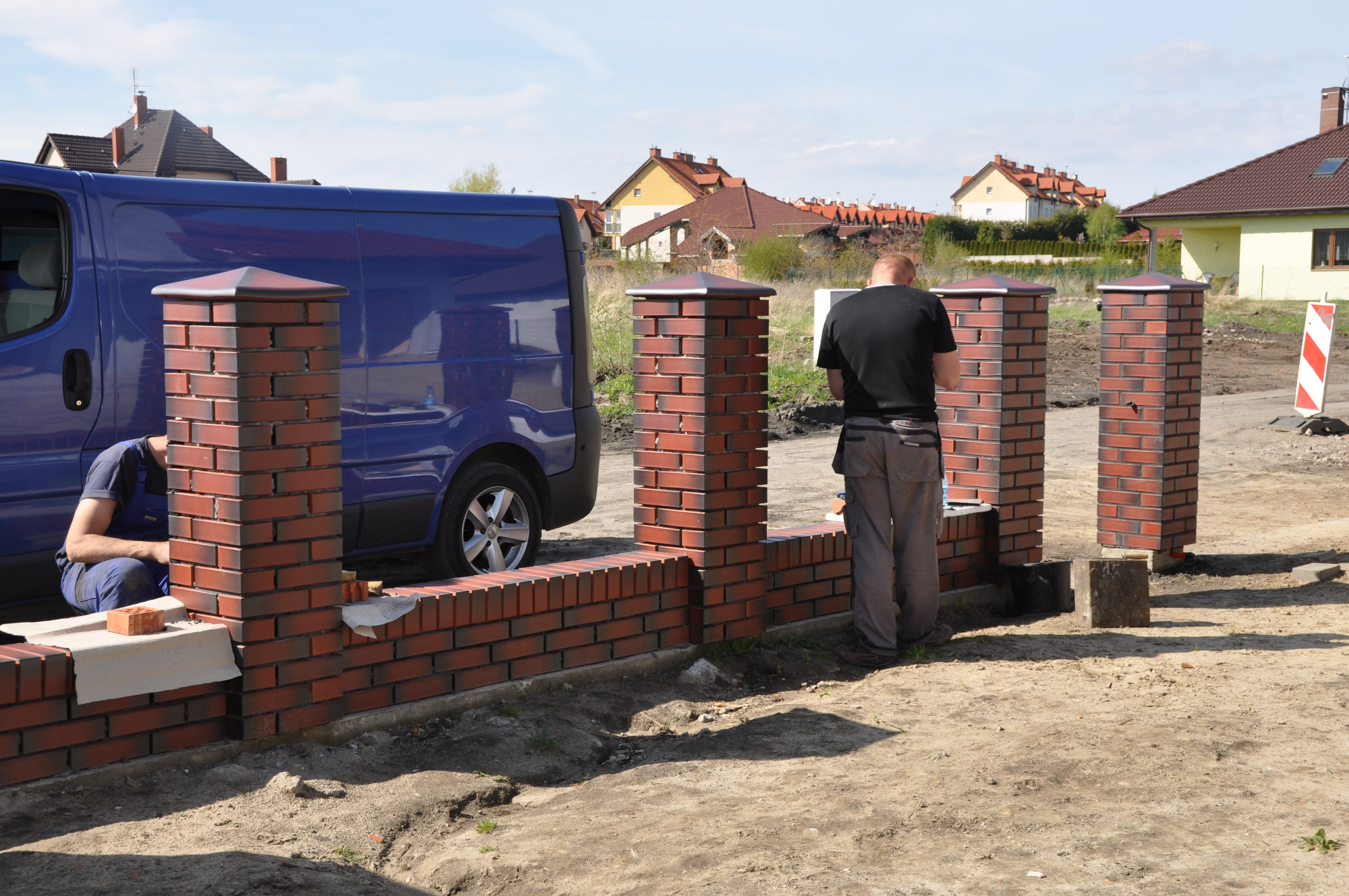 Budowa ogrodzenia z cegły klinkierowej: murarze w trakcie pracy, widoczny fragment muru, słupki i zaparkowany niebieski bus.