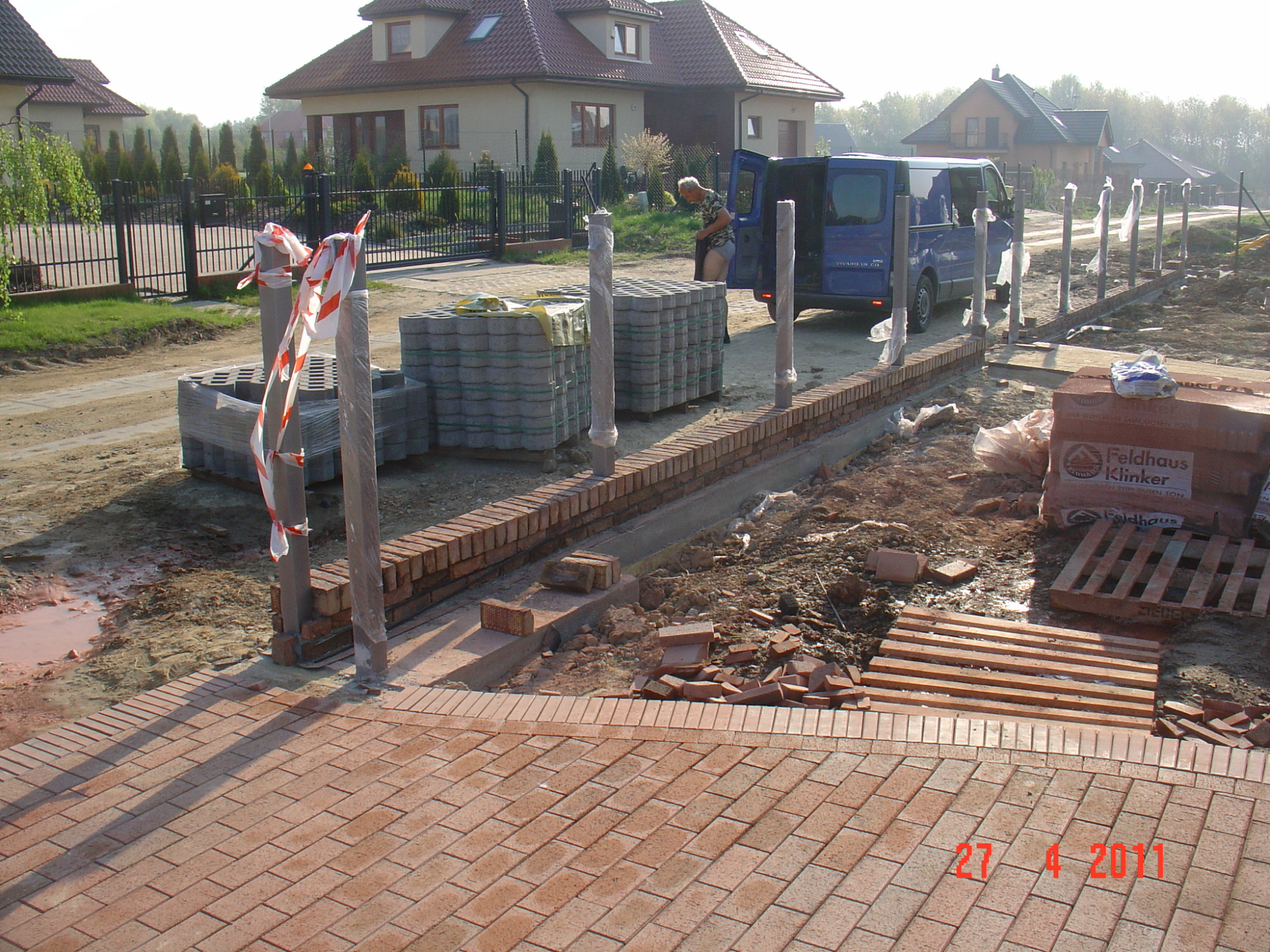 Budowa ogrodzenia z klinkieru i słupków w folii, widoczne palety z cegłami klinkierowymi Feldhaus Klinker, ułożona kostka klinkierowa na podjeździe, oraz niebieski bus z otwartymi drzwiami w tle.