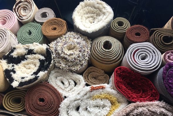 Stos zwiniętych dywanów o różnych wzorach i kolorach, w tym dywan z czarno-białym wzorem, dywan shaggy z fioletowymi akcentami i dywan z czerwonym runem, ułożonych ciasno obok siebie.