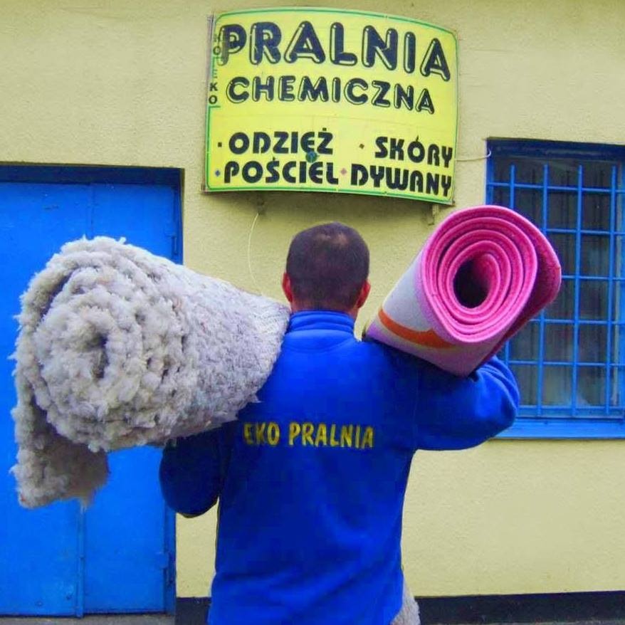 Mężczyzna z tyłu w niebieskiej bluzie z napisem 'EKO PRALNIA' niesie dwa zwinięte dywany, jeden jasny i puszysty, drugi różowo-pomarańczowy, przed budynkiem pralni chemicznej z żółtym szyldem.