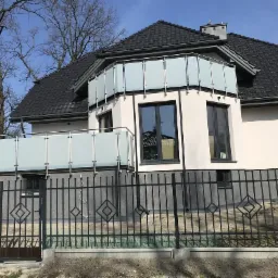 Nowoczesny dom z balkonem zabezpieczonym balustradą szklaną z elementami ze stali nierdzewnej, widoczna również metalowa brama wjazdowa z ozdobnymi elementami.