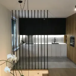 Nowoczesne wnętrze z minimalistyczną kuchnią oddzieloną od schodów czarnymi, pionowymi prętami tworzącymi ażurową przegrodę, z widocznym logo firmy Wood Weld w górnym lewym rogu.