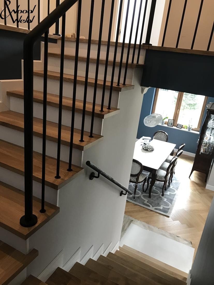Drewniane schody z czarną, metalową balustradą i poręczą, widok z góry na dół, w tle jadalnia z oknem i parkietem w jodełkę.
