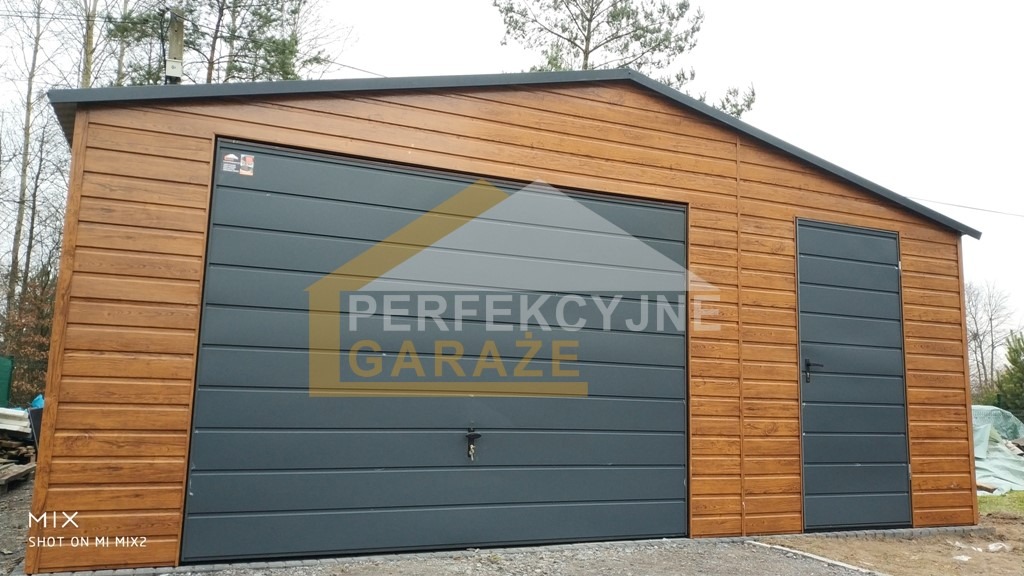 Gotowy garaż blaszany z drewnopodobną elewacją i antracytowymi drzwiami segmentowymi oraz drzwiami wejściowymi, widoczne drzewa w tle.