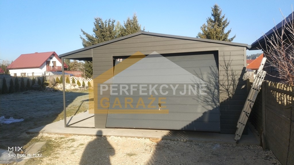 Nowoczesny, grafitowy garaż blaszany z podjazdem, częściowo otwartą bramą segmentową i dobudowanym zadaszeniem, widoczny na tle ogrodu z przymrozkami.