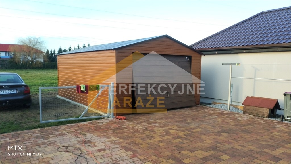 Gotowy, brązowy garaż blaszany z bramą segmentową, widoczny z zewnątrz na posesji z kostką brukową, obok samochód i suszarka na pranie.