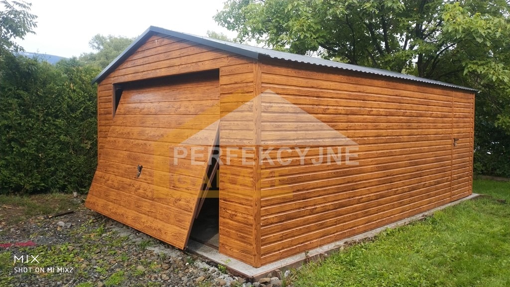 Garaż blaszany z częściowo otwartymi drzwiami przesuwnymi, imitacja drewna, zamek na bocznej ścianie, otoczony zielenią.