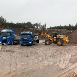 Żółta ładowarka Hyundai załadowuje materiał do dwóch niebieskich ciężarówek Scania na placu budowy z widocznym wyrobiskiem skalnym w tle.