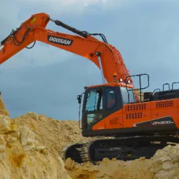 Pomarańczowa koparka Doosan DX300LC z łyżką pełną piasku, pracująca w wykopie o ścianach z jasnych skał wapiennych, na tle pochmurnego nieba.