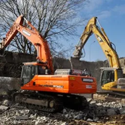 Dwie koparki, pomarańczowa Doosan i żółta CAT, podczas rozbiórki betonowego mostu, z widocznym fragmentem znaku ostrzegawczego i pracownikiem w pomarańczowej kamizelce w tle.