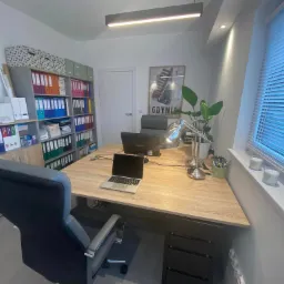 Jasne biuro z dużym biurkiem, laptopem, dwoma monitorami, regałem z segregatorami, plakatem Gdyni i roletami w oknie.