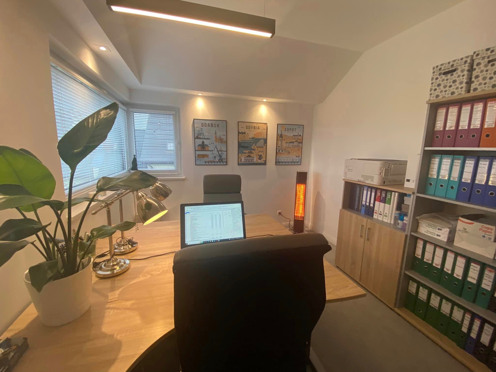 Jasne biuro z biurkiem, laptopem wyświetlającym arkusz kalkulacyjny, lampkami biurkowymi, rośliną doniczkową, regałem z segregatorami i plakatami Gdańska, Gdyni i Sopotu na ścianie.