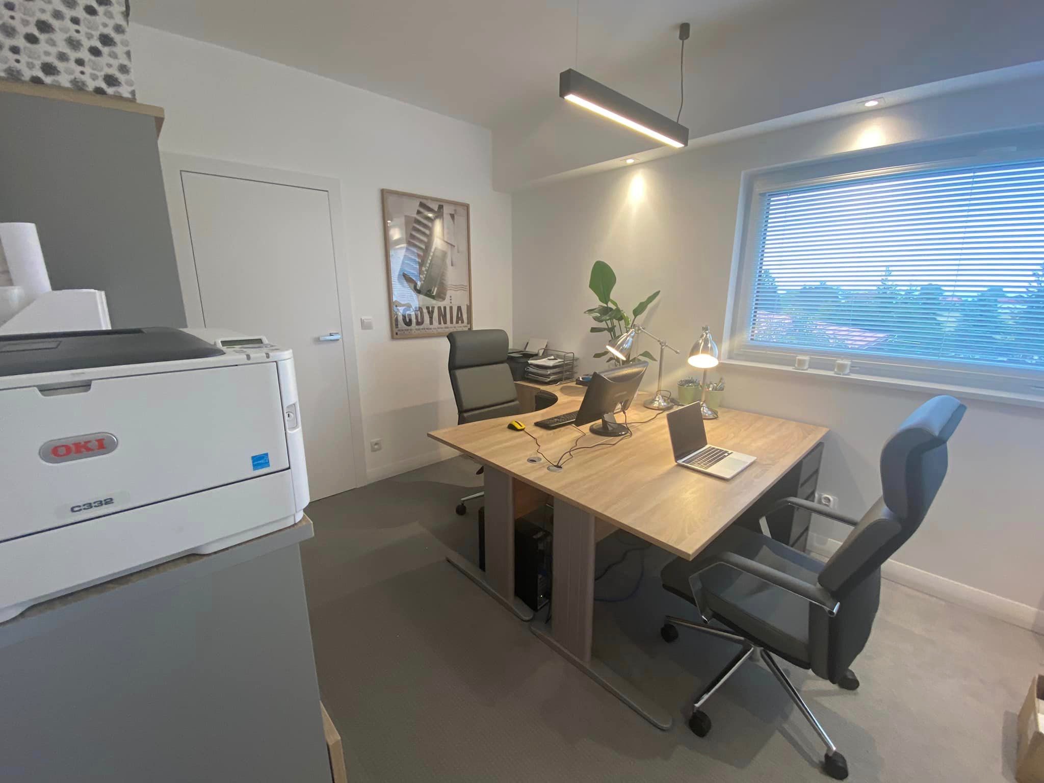 Nowoczesne biuro: jasne drewniane biurko z dwoma stanowiskami komputerowymi, ergonomiczne krzesła, drukarka OKI C332 i plakat Gdyni na ścianie, oświetlone wiszącą lampą LED.