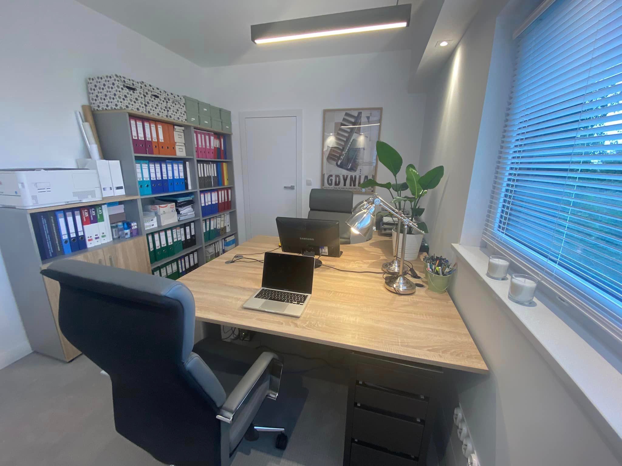 Jasne biuro z dużym biurkiem, laptopem, dwoma monitorami, regałem z segregatorami, plakatem Gdyni i roletami w oknie.