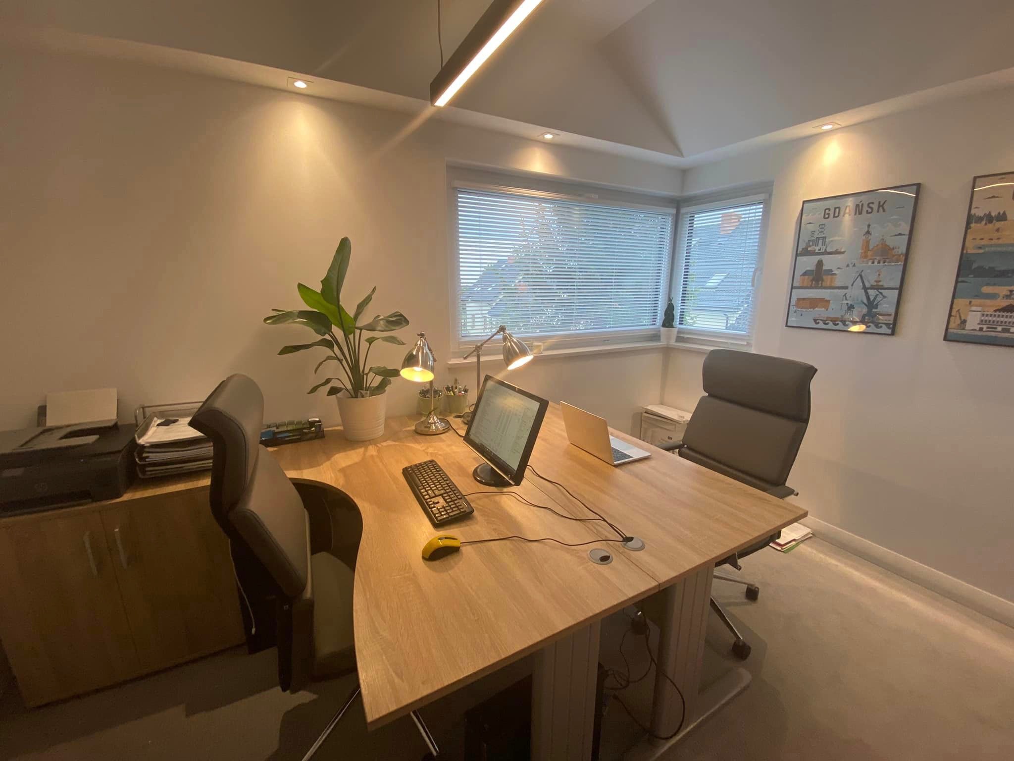 Jasne, nowoczesne biuro z dużym drewnianym biurkiem, komputerem stacjonarnym, laptopem, żółtą myszką, lampkami biurkowymi, rośliną doniczkową i plakatami z motywem Gdańska na ścianie.