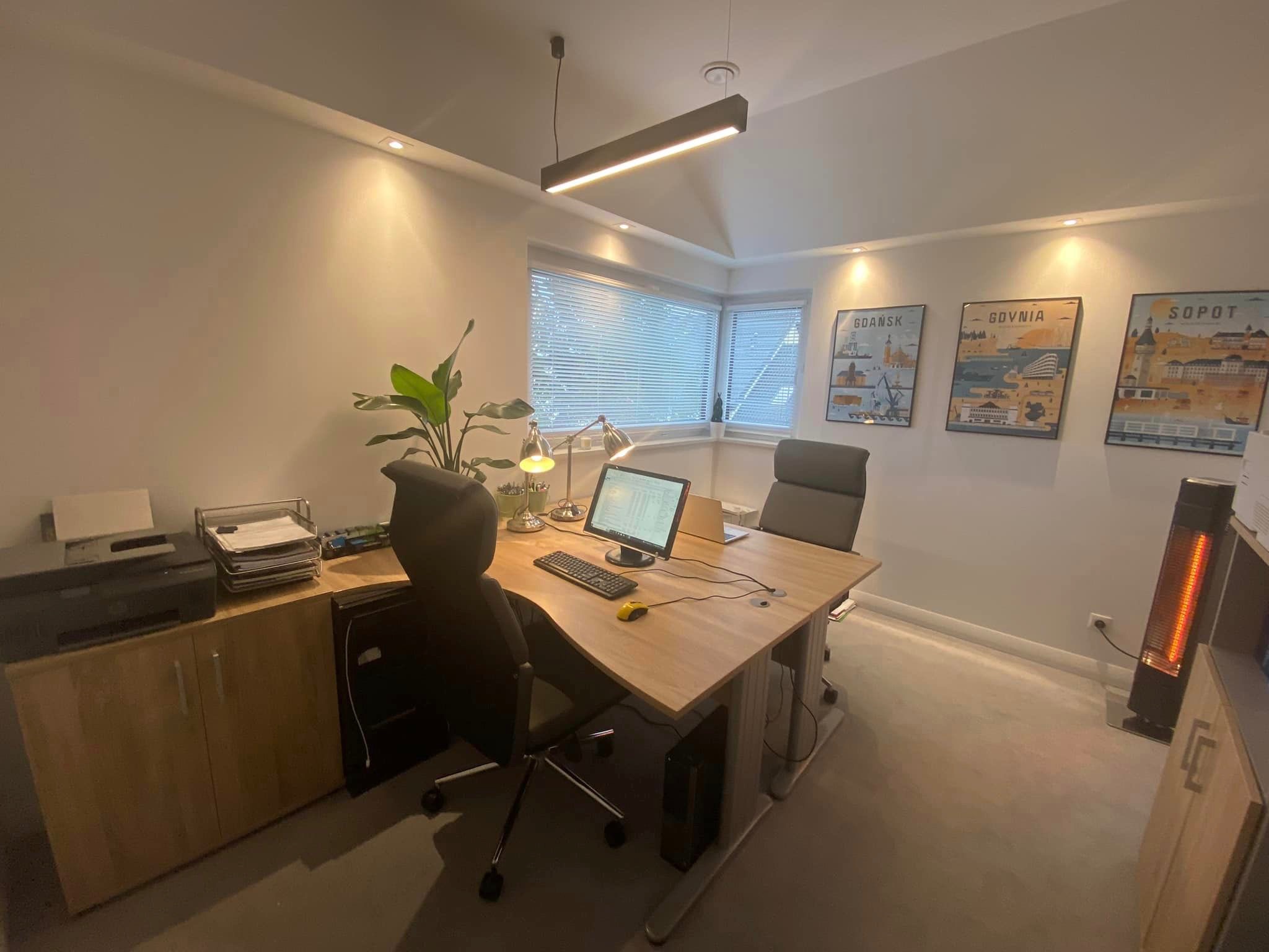 Jasne biuro z drewnianym biurkiem, komputerem, lampką, rośliną doniczkową, drukarką i plakatami miast Gdańsk, Gdynia, Sopot.
