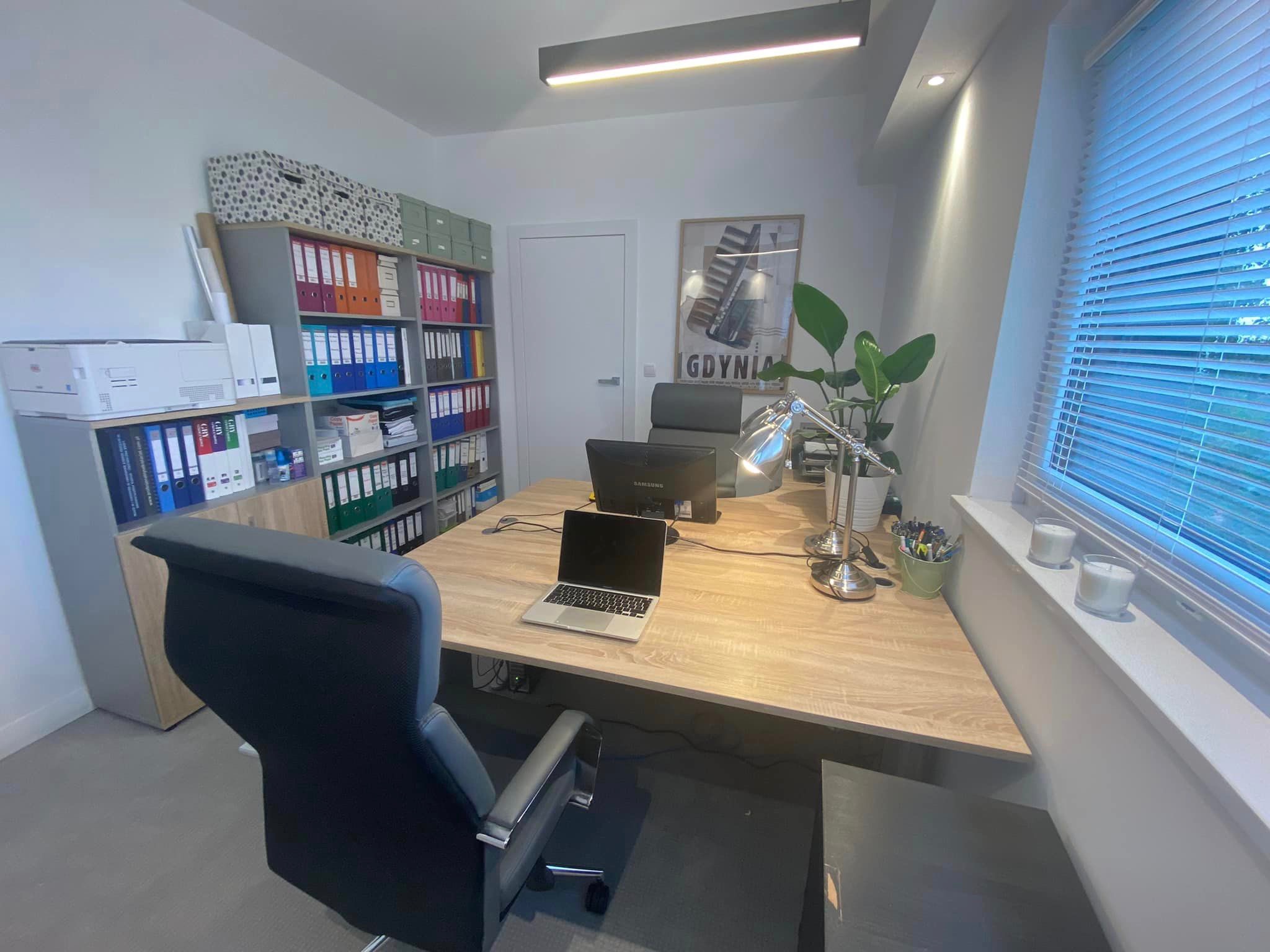 Jasne biuro z dużym biurkiem, laptopem, regałem pełnym segregatorów, plakatem 'Gdynia' na ścianie i rośliną doniczkową przy oknie.