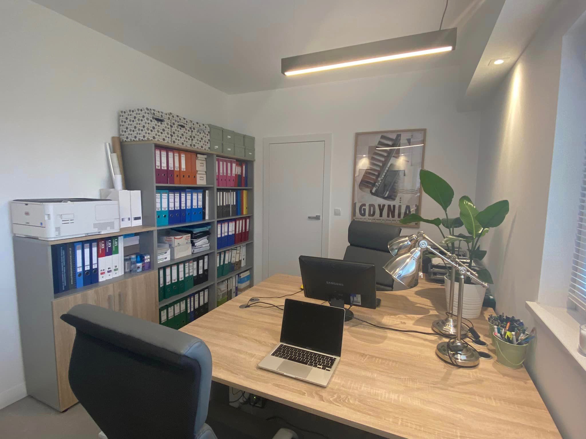Jasne biuro z biurkiem z laptopem, dwoma monitorami, lampką, regałem z segregatorami i plakatem Gdyni na ścianie.