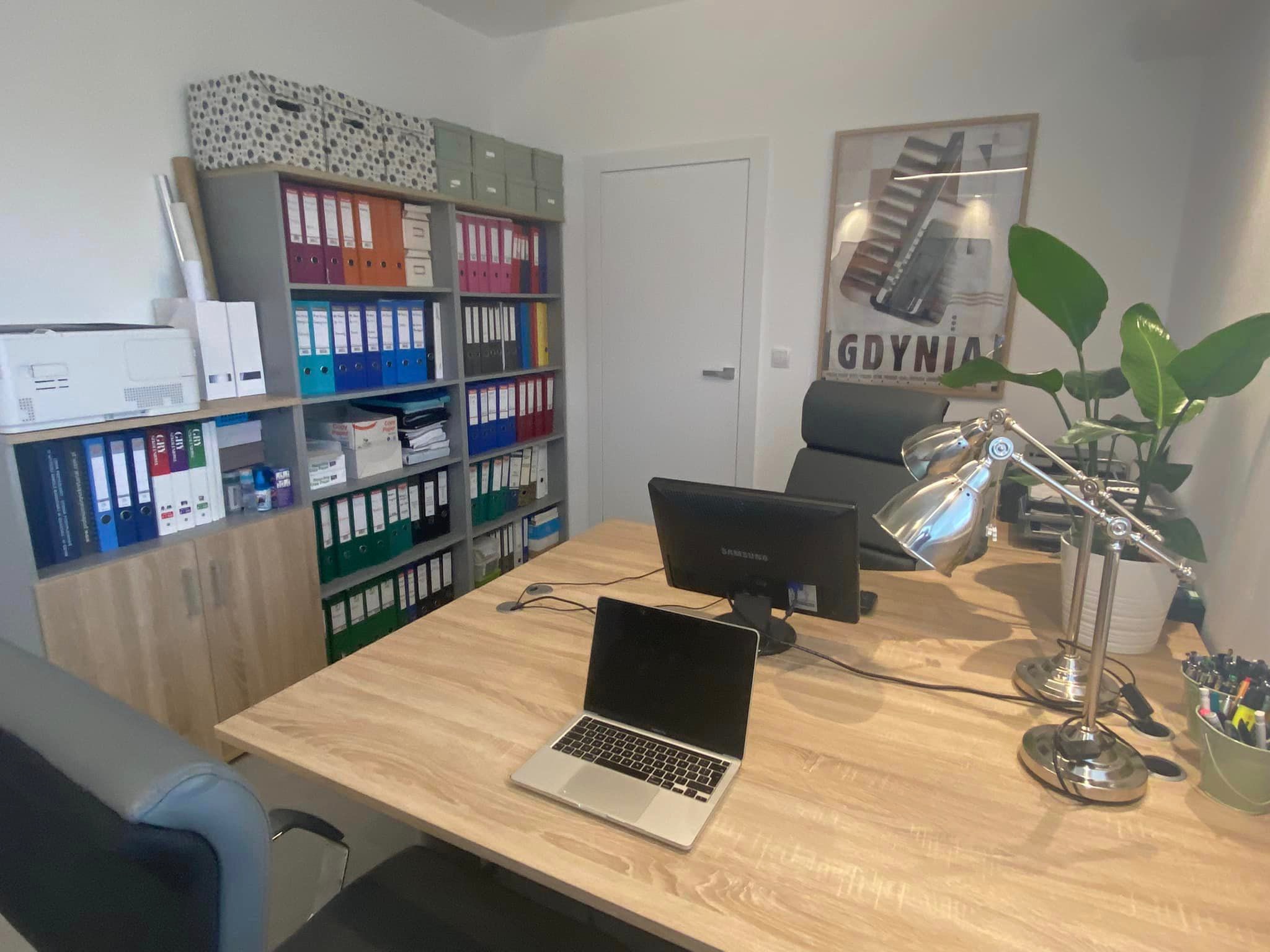 Biuro z drewnianym biurkiem, laptopem, monitorem, lampką, regałem z segregatorami, drukarką i plakatem Gdyni na ścianie.