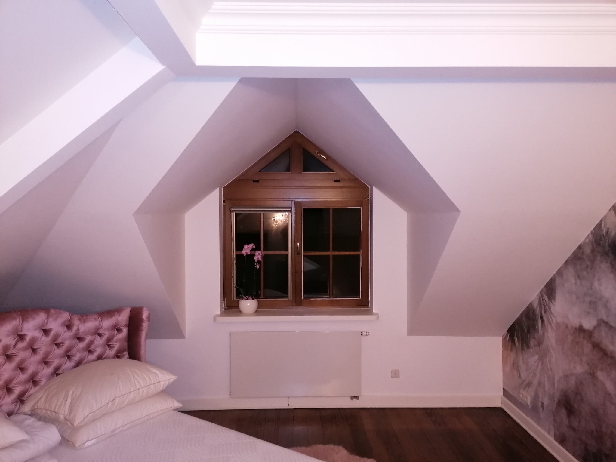 Pokój na poddaszu z drewnianym oknem w kształcie trójkąta, różowa orchidea w doniczce na parapecie, biały grzejnik panelowy, ściana z wzorzystą tapetą, łóżko z pikowanym zagłówkiem.