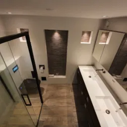 Nowoczesna łazienka z kabiną prysznicową typu walk-in z czarnymi ramami, podwójną umywalką nablatową z ciemnymi szafkami, drewnopodobną podłogą, wnękami z dekoracyjną mozaiką i oświetleniem punktowym.