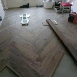 Układanie parkietu w jodełkę z widocznymi krzyżykami dystansowymi, fragment surowego drewna i okna balkonowe w tle.