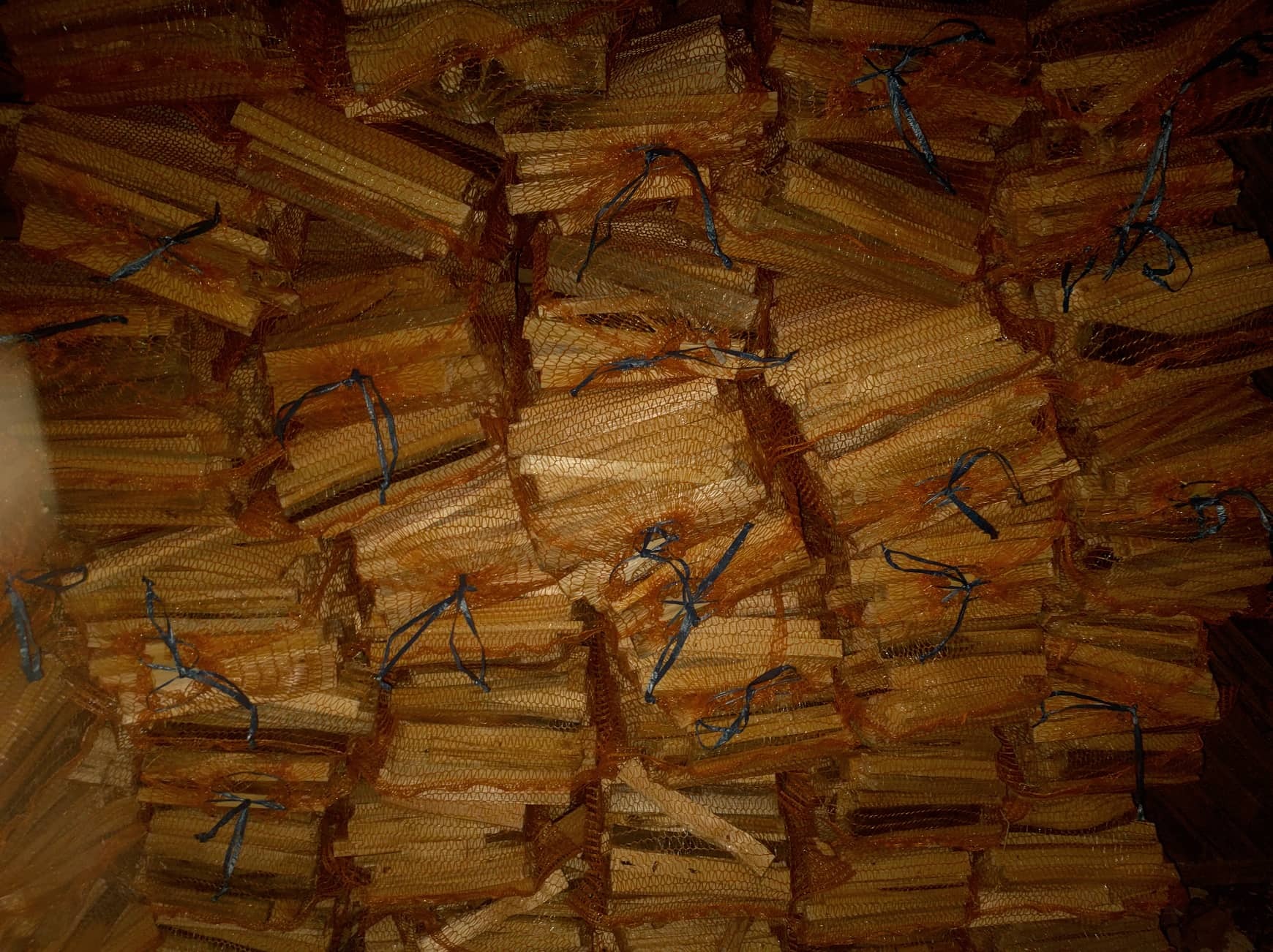 Stos worków siatkowych wypełnionych pociętymi kawałkami drewna, związanych niebieskim sznurkiem, tworzących uporządkowany, ale surowy magazyn opału.