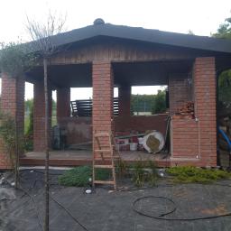 Ar3i bud Artur Kurdziel - Murowana altana w trakcie budowy, widoczne cegły, drabina, materiały izolacyjne i narzędzia na drewnianej podłodze oraz przygotowane rośliny do nasadzeń wokół.