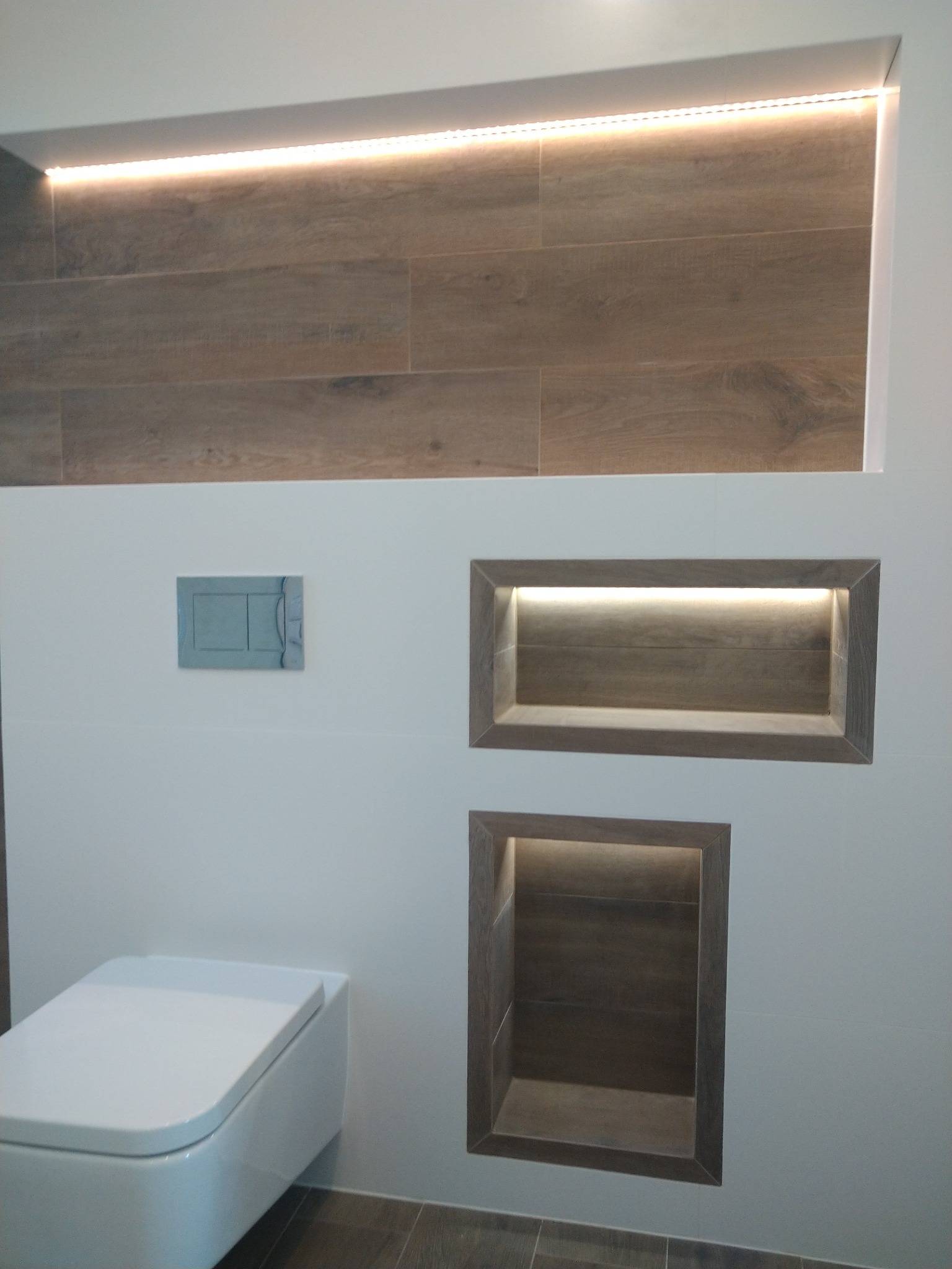 Nowoczesna łazienka z podwieszaną toaletą, wnękami z podświetleniem LED wykończonymi płytkami imitującymi drewno oraz płytkami podłogowymi w tym samym stylu.