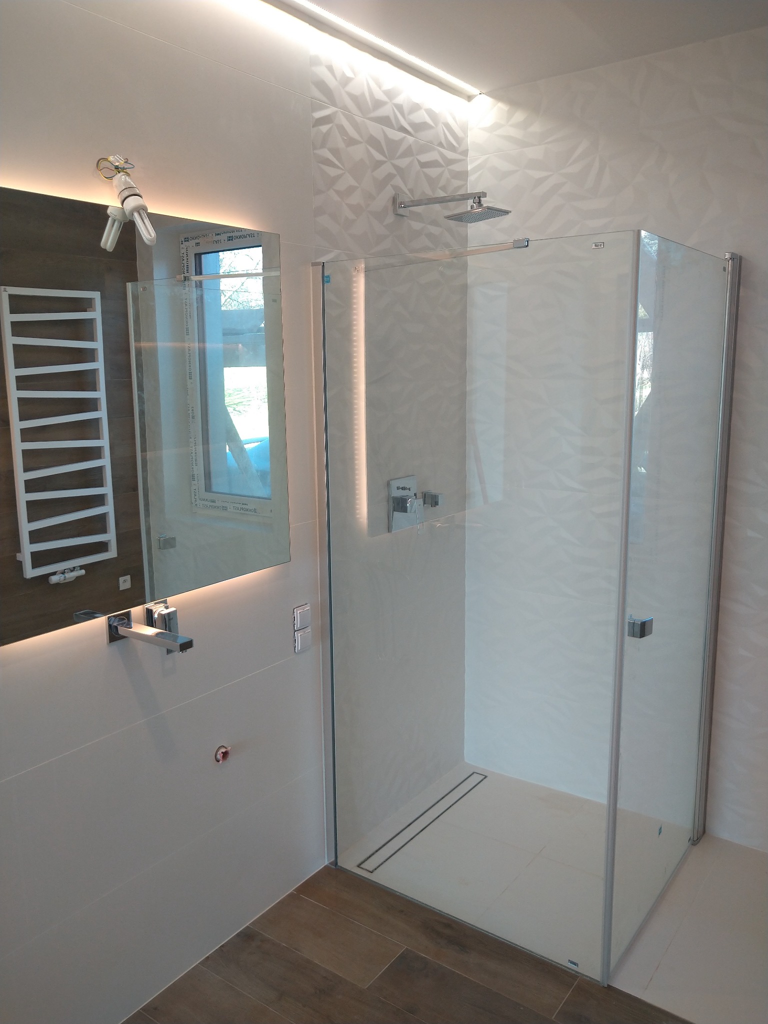 Nowoczesna łazienka z kabiną prysznicową typu walk-in, armaturą w kolorze chromu i podświetlanym lustrem z grzejnikiem drabinkowym w odbiciu.