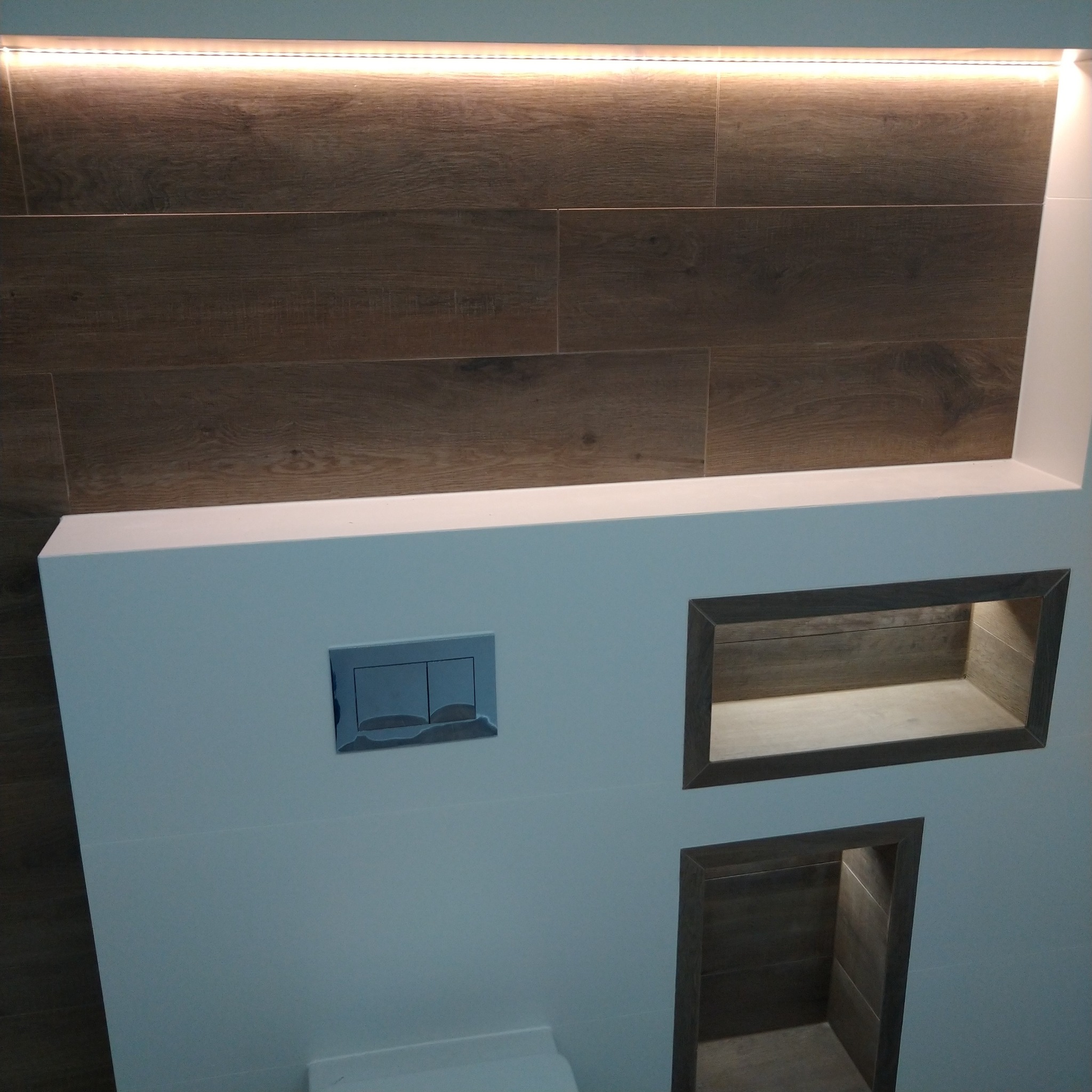 Nowoczesna łazienka z drewnopodobnymi płytkami, podświetleniem LED nad górną krawędzią, wnękami w ścianie z drewnopodobnym wykończeniem i chromowanym przyciskiem spłuczki.