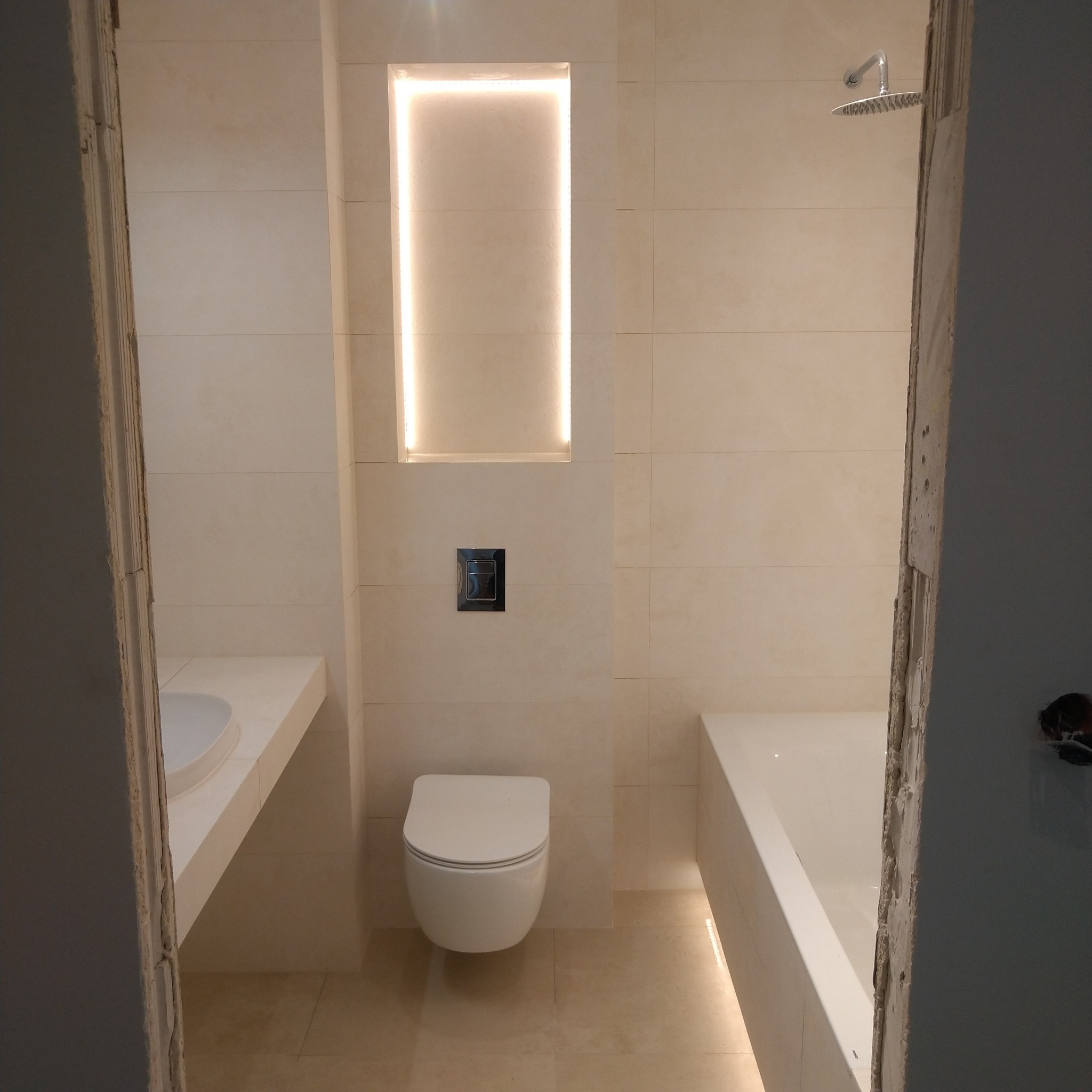 Wykończona łazienka w jasnych beżowych płytkach z podświetloną wnęką, wiszącą toaletą i umywalką w blacie, widok zza futryny w trakcie remontu.